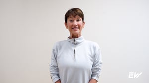 EW personal training client stands against a white wall and wears a white long sleeve shirt to talk about her experience and decreased shoulder pain. 