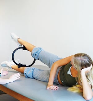 Physical therapist guiding a dancer through a side-lying hip abduction exercise using a Pilates ring for hip strength and turnout rehabilitation on a treatment table at a physical therapy clinic.