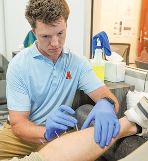 Licensed physical therapist performing dry needling on a patient's lower leg muscle trigger point using a thin filiform needle for pain relief and muscle tension release at a physical therapy clinic.
