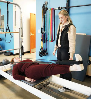 Pilates instructor supervising client performing reformer leg strap exercise for core and lower back pain rehabilitation.