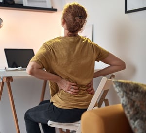 Woman sitting at a desk holding her lower back, illustrating office-related low back pain and poor sitting posture, commonly treated with physical therapy for spinal alignment, core strengthening, and pain relief.