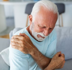 Older man experiencing shoulder pain at home, holding his shoulder due to age-related joint discomfort commonly treated with physical therapy.