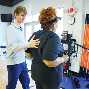 Physical therapist providing hands-on shoulder guidance to a patient performing cable machine exercises for shoulder rehabilitation in a modern physical therapy gym.