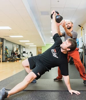 Personal trainer spotting man performing side plank kettlebell press at EW Motion Therapy.