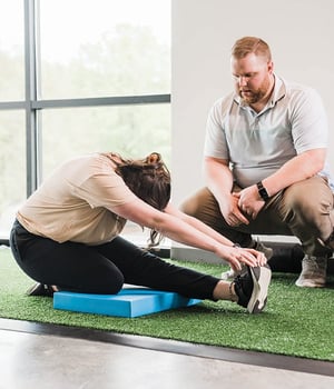 Personal trainer observing woman perform seated hamstring stretch on balance pad at EW Motion Therapy.