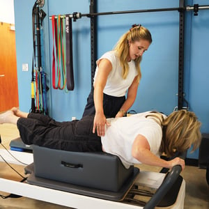 One-on-one clinical Pilates session with an instructor manually guiding a patient's lower back alignment during a prone reformer exercise for spinal rehabilitation.