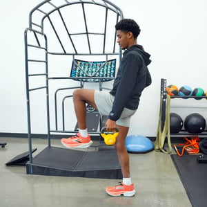 Young athlete performing single-leg kettlebell exercise for sports recovery and injury rehabilitation at a physical therapy clinic.