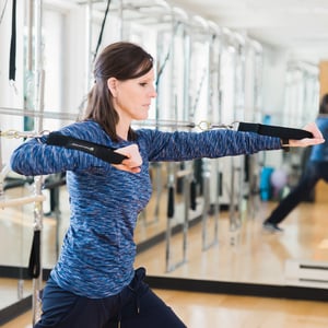 Pilates tower wall unit exercise for functional strength and postural rehabilitation showing a patient in a warrior stance using spring resistance for upper body and hip training.