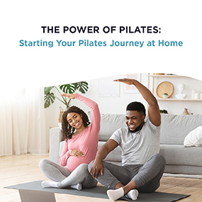 Pregnant woman and her partner sitting on a yoga mat doing side stretches together at home, representing EW Motion Therapy's guide to starting your Pilates journey at home